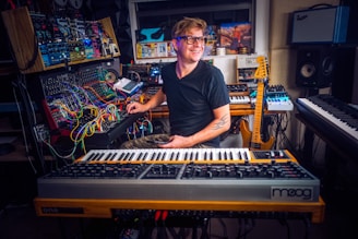 A person is sitting in a home studio surrounded by various musical instruments and equipment. The setup includes synthesizers, a mixing console, and a guitar. The person appears to be focusing on a synthesizer while smiling pleasantly, creating a creative and enthusiastic atmosphere.