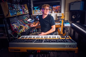 A person is sitting in a home studio surrounded by various musical instruments and equipment. The setup includes synthesizers, a mixing console, and a guitar. The person appears to be focusing on a synthesizer while smiling pleasantly, creating a creative and enthusiastic atmosphere.