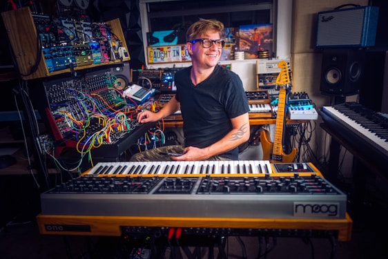 A person is sitting in a home studio surrounded by various musical instruments and equipment. The setup includes synthesizers, a mixing console, and a guitar. The person appears to be focusing on a synthesizer while smiling pleasantly, creating a creative and enthusiastic atmosphere.