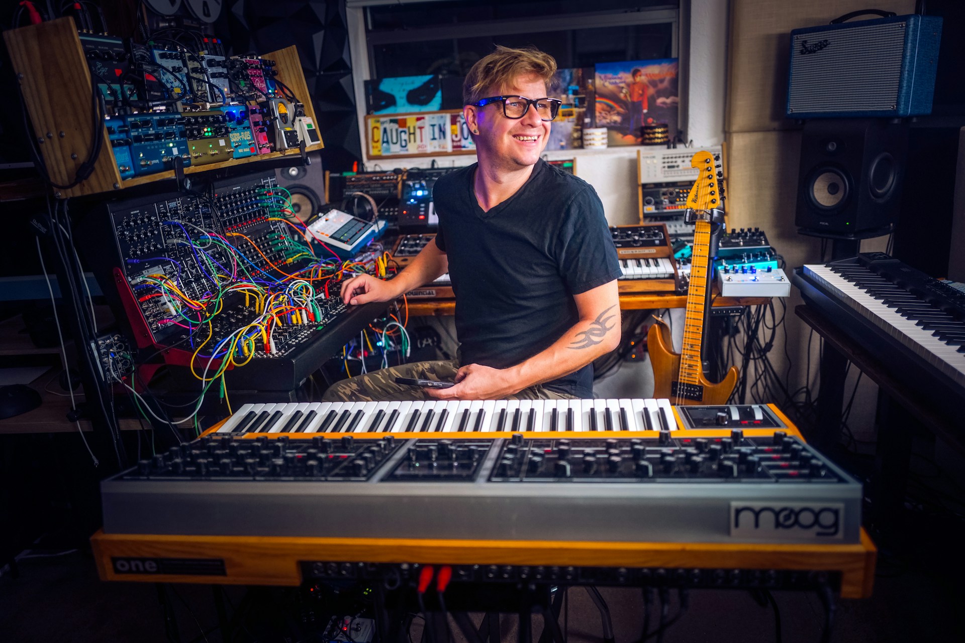 A person is sitting in a home studio surrounded by various musical instruments and equipment. The setup includes synthesizers, a mixing console, and a guitar. The person appears to be focusing on a synthesizer while smiling pleasantly, creating a creative and enthusiastic atmosphere.