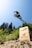 A mountain biker is performing a high jump off a ramp in an outdoor setting. The biker is captured mid-air against a backdrop of tall trees and a clear blue sky. A wooden ramp with signage, possibly indicating a bike park or event, is visible in the image.