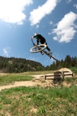A person is performing an aerial stunt on a mountain bike against a backdrop of a clear blue sky with scattered clouds. They are wearing protective gear, including a helmet, and are high above the ground, highlighting the skill and thrill of the action. The landscape below features a dirt path, green grassy area, and a forested hillside.