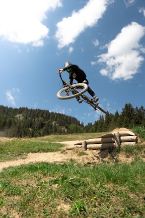 A person is performing an aerial stunt on a mountain bike against a backdrop of a clear blue sky with scattered clouds. They are wearing protective gear, including a helmet, and are high above the ground, highlighting the skill and thrill of the action. The landscape below features a dirt path, green grassy area, and a forested hillside.