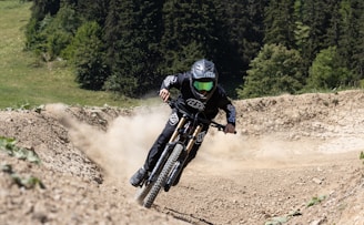 A mountain biker is racing down a dirt trail, kicking up dust as they maneuver through a curve. The rider wears protective gear, including a helmet with a visor and gloves, and the bike is equipped with heavy-duty tires for rough terrain. Surrounding the path are trees and patches of green grass, indicating a forested hillside environment.