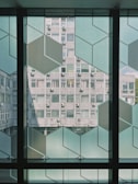 The image displays a modern architectural scene with a view through a decorative glass panel featuring geometric patterns. In the background, there is a concrete building with multiple windows and air conditioning units attached. The photo is taken from the interior of a building, with light filtering through the large windows.
