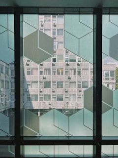 The image displays a modern architectural scene with a view through a decorative glass panel featuring geometric patterns. In the background, there is a concrete building with multiple windows and air conditioning units attached. The photo is taken from the interior of a building, with light filtering through the large windows.