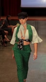 A person is walking down a runway dressed in a stylish outfit featuring a green vest and pants set with an intricate pattern. A white blouse with flared sleeves is worn underneath, paired with a neck scarf and a black cap.