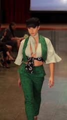 A person is walking down a runway dressed in a stylish outfit featuring a green vest and pants set with an intricate pattern. A white blouse with flared sleeves is worn underneath, paired with a neck scarf and a black cap.