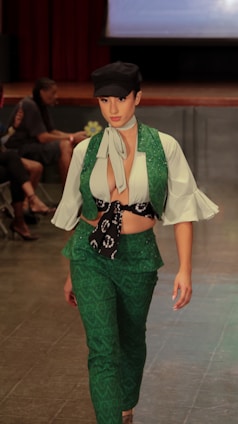 A person is walking down a runway dressed in a stylish outfit featuring a green vest and pants set with an intricate pattern. A white blouse with flared sleeves is worn underneath, paired with a neck scarf and a black cap.
