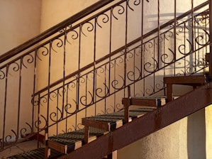 A metal staircase with ornate, swirling patterns on the railing. The lighting casts soft shadows, creating an artistic interplay of lines on the wall.