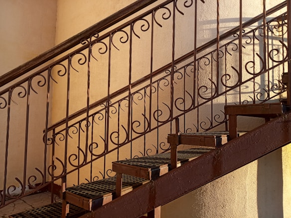 A metal staircase with ornate, swirling patterns on the railing. The lighting casts soft shadows, creating an artistic interplay of lines on the wall.