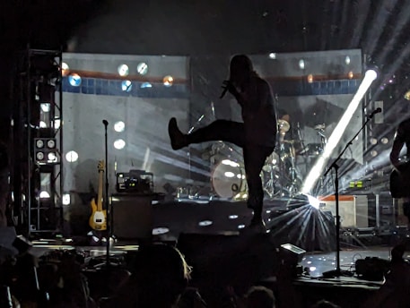 A silhouetted performer on stage is dramatically lit by bright lights, creating a dynamic scene. In the background, various musical equipment and instruments are visible including a guitar and drums. The atmosphere suggests a lively music concert.