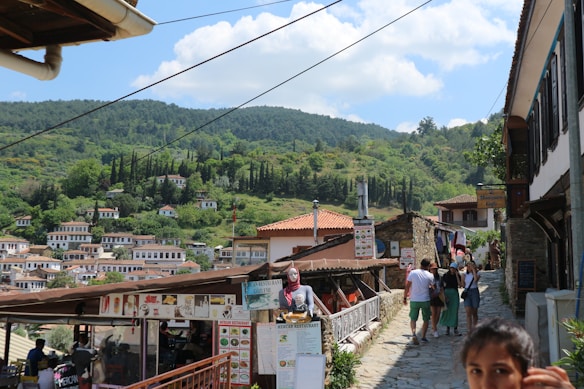 A charming village scene with traditional houses featuring red-tiled roofs nestled on a hillside. A bustling pathway with people walking by is lined with small shops and cafes, adorned with colorful signs and awnings. Lush greenery and trees fill the background under a partly cloudy blue sky.