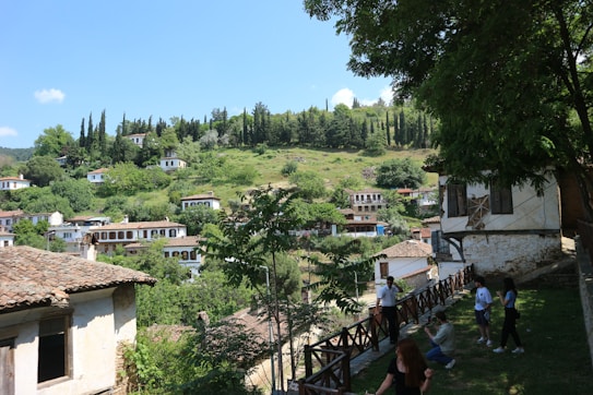 A scenic hillside village with traditional whitewashed houses featuring red-tiled roofs, surrounded by lush greenery and tall trees under a bright blue sky. People are seen walking along a wooden fence, enjoying the natural landscape.