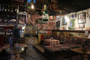 A cozy, rustic interior with wooden furniture and an eclectic mix of decorations. There are wine bottles on shelves, vintage posters on the walls, and traditional patterned fabric on the seating and table. The space is dimly lit by hanging light bulbs, creating a warm and inviting atmosphere. Various objects such as a dartboard, pottery, and framed pictures add to the charm.