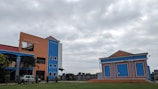 A brightly colored building with orange and blue walls stands prominently under a cloudy sky. There is an adjacent smaller building with a similar color scheme. Several windows are visible, along with a sign that reads 'Madhusudan Gardens'. The surrounding area has some greenery and a vehicle is parked near the main building.