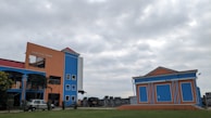 A brightly colored building with orange and blue walls stands prominently under a cloudy sky. There is an adjacent smaller building with a similar color scheme. Several windows are visible, along with a sign that reads 'Madhusudan Gardens'. The surrounding area has some greenery and a vehicle is parked near the main building.