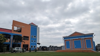 A brightly colored building with orange and blue walls stands prominently under a cloudy sky. There is an adjacent smaller building with a similar color scheme. Several windows are visible, along with a sign that reads 'Madhusudan Gardens'. The surrounding area has some greenery and a vehicle is parked near the main building.