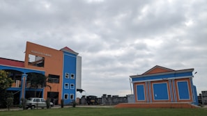 A brightly colored building with orange and blue walls stands prominently under a cloudy sky. There is an adjacent smaller building with a similar color scheme. Several windows are visible, along with a sign that reads 'Madhusudan Gardens'. The surrounding area has some greenery and a vehicle is parked near the main building.