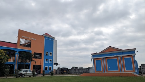 A brightly colored building with orange and blue walls stands prominently under a cloudy sky. There is an adjacent smaller building with a similar color scheme. Several windows are visible, along with a sign that reads 'Madhusudan Gardens'. The surrounding area has some greenery and a vehicle is parked near the main building.