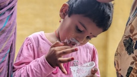 A young child wearing a pink shirt is engrossed in eating something creamy, with a dollop on their nose and mouth. The child holds a small, clear cup in one hand and has creamy fingers. Two adults are partially visible on either side, perhaps helping or watching the child.