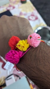 A wrist adorned with a handmade bracelet featuring colorful pom-poms in shades of red, yellow, pink, and magenta. The bracelet is tied around the wrist against a background that appears to be indoors with a table or cloth in muted tones.