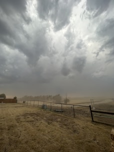 Dark storm clouds gather ominously in the sky above a barren field with dry grass. A metal fence runs through the foreground, and trees are visible in the hazy distance. The atmosphere is heavy, suggesting an impending storm.