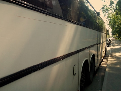 A large white coach bus parked on the side of a street on a sunny day, with trees casting shadows on the pavement and part of the vehicle. The bus has tinted windows and is in close proximity to other parked cars along the street.