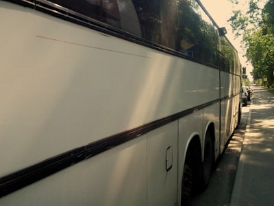 A large white coach bus parked on the side of a street on a sunny day, with trees casting shadows on the pavement and part of the vehicle. The bus has tinted windows and is in close proximity to other parked cars along the street.