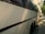 A large white coach bus parked on the side of a street on a sunny day, with trees casting shadows on the pavement and part of the vehicle. The bus has tinted windows and is in close proximity to other parked cars along the street.