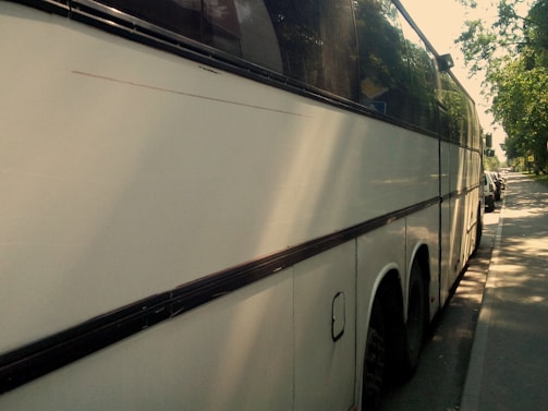 A large white coach bus parked on the side of a street on a sunny day, with trees casting shadows on the pavement and part of the vehicle. The bus has tinted windows and is in close proximity to other parked cars along the street.