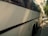 A large white coach bus parked on the side of a street on a sunny day, with trees casting shadows on the pavement and part of the vehicle. The bus has tinted windows and is in close proximity to other parked cars along the street.