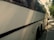 A large white coach bus parked on the side of a street on a sunny day, with trees casting shadows on the pavement and part of the vehicle. The bus has tinted windows and is in close proximity to other parked cars along the street.