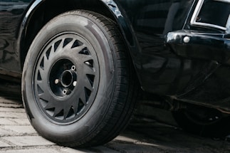 A close-up view of a car's front wheel and tire on a cobblestone surface. The wheel features a detailed rim design, and the car's black paint has a glossy finish.