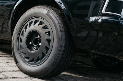 A close-up view of a car's front wheel and tire on a cobblestone surface. The wheel features a detailed rim design, and the car's black paint has a glossy finish.