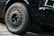 A close-up view of a car's front wheel and tire on a cobblestone surface. The wheel features a detailed rim design, and the car's black paint has a glossy finish.
