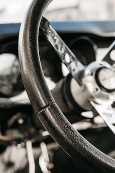 Close-up of a vintage Formula 1 steering wheel with worn leather grips and polished metal spokes, set against a dark racing cockpit background.
