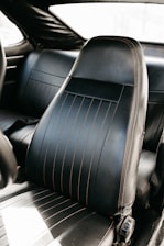 A close-up view of a black leather car seat with vertical stitching and slight signs of wear. The seat is positioned inside the car, with a glimpse of the backseat visible.