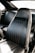 A close-up view of a black leather car seat with vertical stitching and slight signs of wear. The seat is positioned inside the car, with a glimpse of the backseat visible.