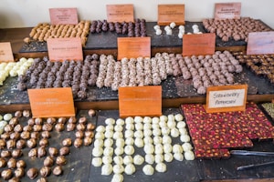 An assortment of chocolates and truffles is displayed on tiered shelves. Each section is labeled with small wooden signs indicating the flavor, such as 'Popping Candy' and 'Irish Whiskey Cream.' The chocolates vary in appearance, including various shapes and colors, ranging from dark brown to creamy white.