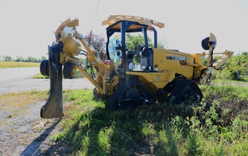 A large, yellow construction vehicle with tracks is situated on an area with gravel and grass. The vehicle features an arm with a large attachment at the front, designed for digging or moving earth. It appears to be an excavator or similar heavy machinery, positioned in a natural setting with fields and some trees in the background.