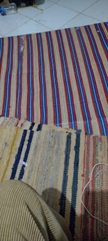 The image features two woven rugs with striped patterns laid out on a tiled floor. One rug has red, blue, and tan stripes, while the other showcases a variety of colors, including yellow, black, blue, and red. In the upper left corner, there is a partially visible water bottle and some debris on the tiles. A piece of clothing or fabric in a striped pattern protrudes into the frame from the bottom left corner.