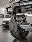 Close-up view of a rugged vehicle's front section in a showroom, focusing on the grille, tire, and towing hitch. The background features another car with visible details in a well-lit environment.