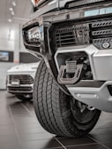 Close-up view of a rugged vehicle's front section in a showroom, focusing on the grille, tire, and towing hitch. The background features another car with visible details in a well-lit environment.