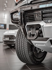 Close-up view of a rugged vehicle's front section in a showroom, focusing on the grille, tire, and towing hitch. The background features another car with visible details in a well-lit environment.
