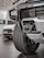 Close-up view of a rugged vehicle's front section in a showroom, focusing on the grille, tire, and towing hitch. The background features another car with visible details in a well-lit environment.