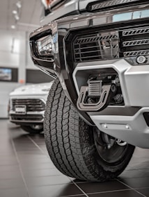 Close-up view of a rugged vehicle's front section in a showroom, focusing on the grille, tire, and towing hitch. The background features another car with visible details in a well-lit environment.
