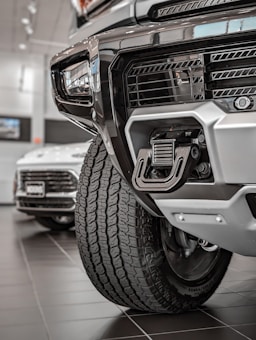 Close-up view of a rugged vehicle's front section in a showroom, focusing on the grille, tire, and towing hitch. The background features another car with visible details in a well-lit environment.