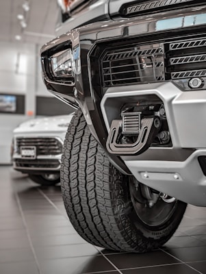 Close-up view of a rugged vehicle's front section in a showroom, focusing on the grille, tire, and towing hitch. The background features another car with visible details in a well-lit environment.