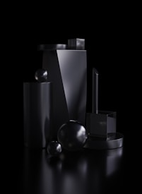 A collection of glossy, black geometric shapes including spheres, cylinders, and cuboids arranged artistically against a dark background. The surfaces of the objects are highly reflective, creating subtle highlights and shadows.
